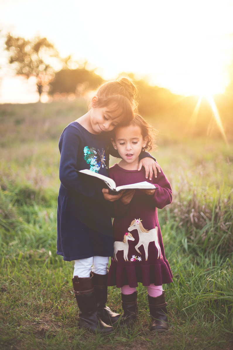 Two little girls reading a book in a field at sunset.